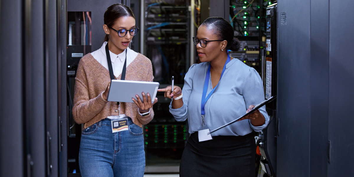 Two women standing near networking devices talking while looking at a laptop